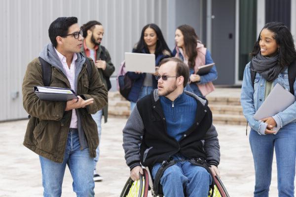 A multiracial group of young adults, university students, hanging out outside a campus building, conversing. The friends include a man with cerebral palsy in a wheelchair. Three of the students are walking and conversing in the foreground while three others are out of focus in the background.